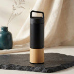 A bamboo and stainless steel thermos sits on a charcoal stone surface next to a teal vase in a bright, minimalist room.